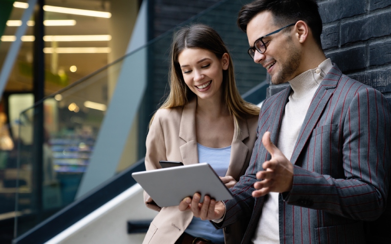 A man and woman stand smiling in a modern office, discussing and pointing at a tablet. The setting conveys a collaborative and professional atmosphere.