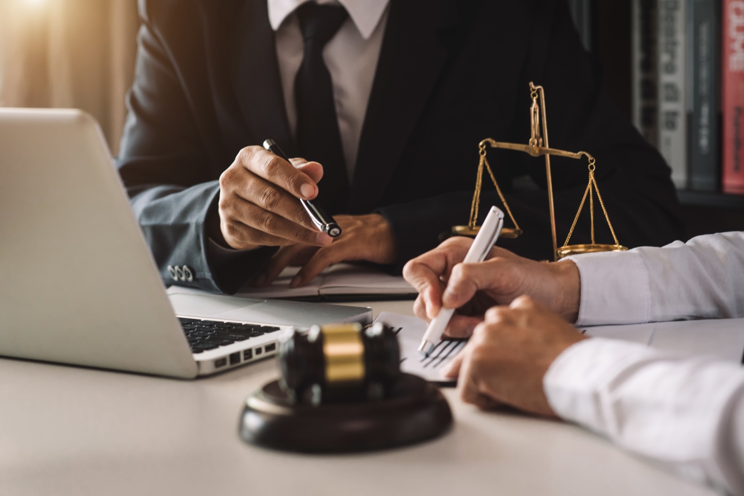 Two professionals in suits discuss legal matters at a desk with a laptop, taking notes, and a golden scale of justice in the background.