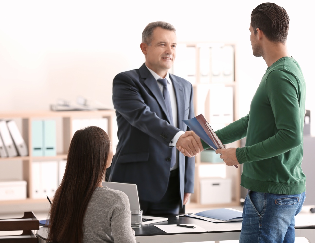 A man in a suit and a man in a green sweater shake hands in an office, while a woman seated at a desk observes. The mood is professional and friendly.
