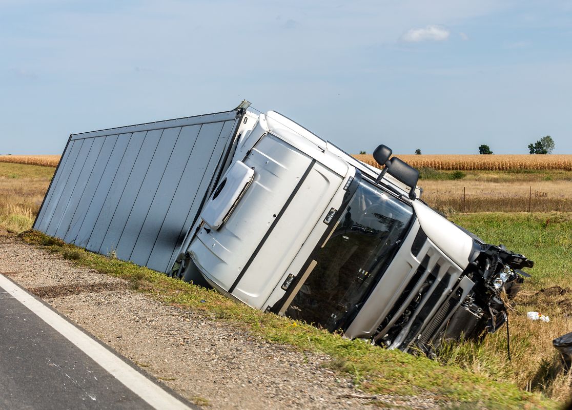 A white truck lies on its side in a grassy ditch beside a rural road, with fields visible in the background under a clear blue sky.