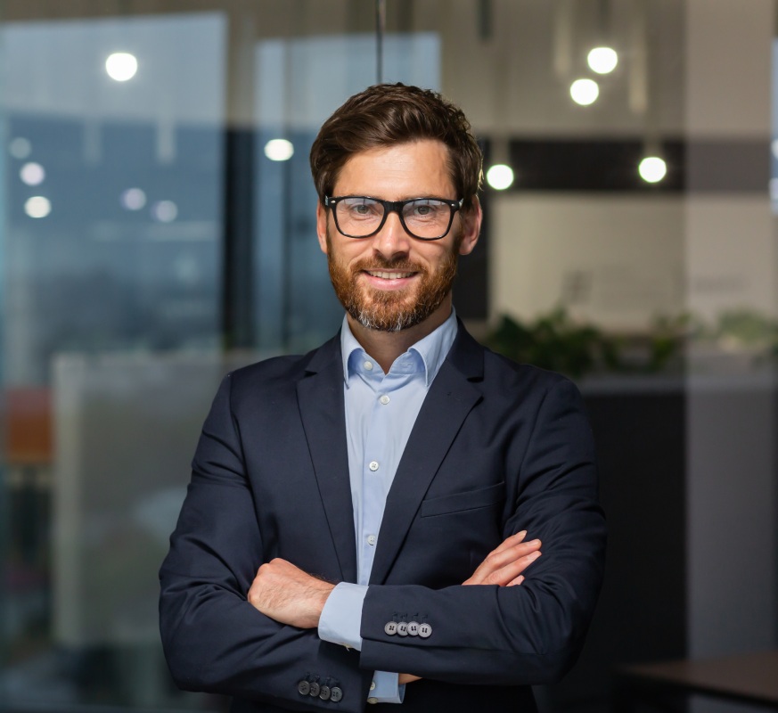 Portrait of successful mature boss, senior businessman in glasses and business suit looking at camera and smiling, man with crossed arms working inside modern office building.