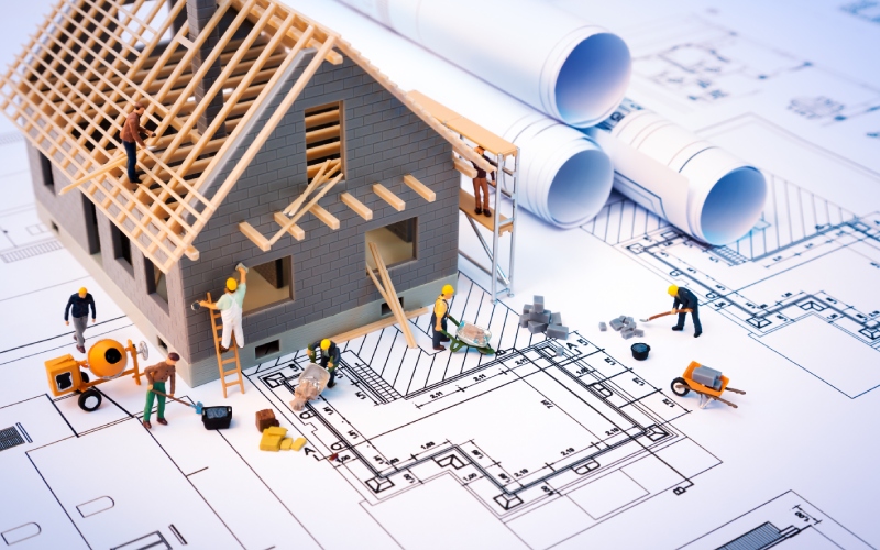 Miniature construction scene with a model house being built on architectural plans. Tiny figures work with tools and blueprints, conveying a busy planning atmosphere.