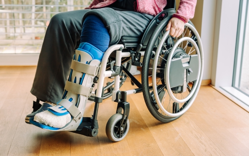 A person in a wheelchair with a leg cast is sitting indoors near a window. The atmosphere is calm, emphasizing recovery and mobility aids.