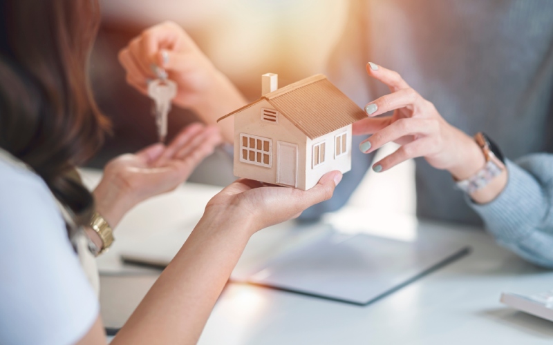 Two people exchange keys and a small house model, symbolizing a home purchase. The scene is warm and bright, conveying a sense of accomplishment.
