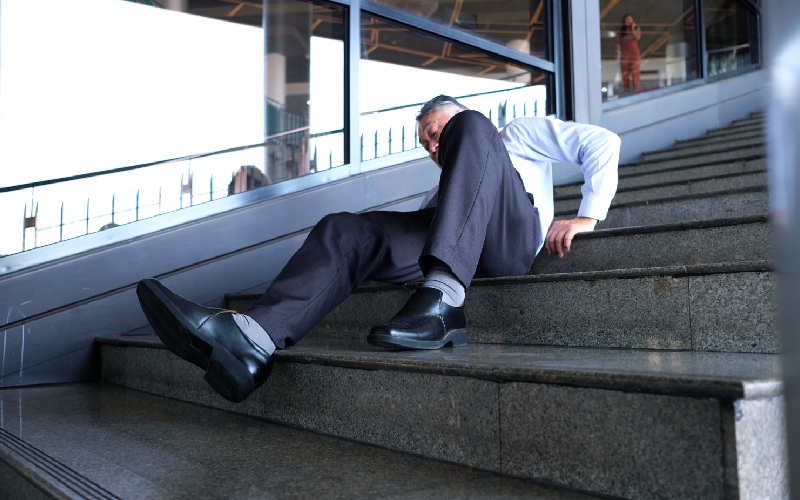 A person in business attire has fallen on a set of concrete stairs, appearing dazed. The indoor setting adds a sense of urgency and concern.