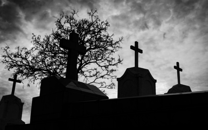 Silhouetted graves with crosses against a moody, cloudy sky and leafless tree. The scene conveys a somber, eerie atmosphere.